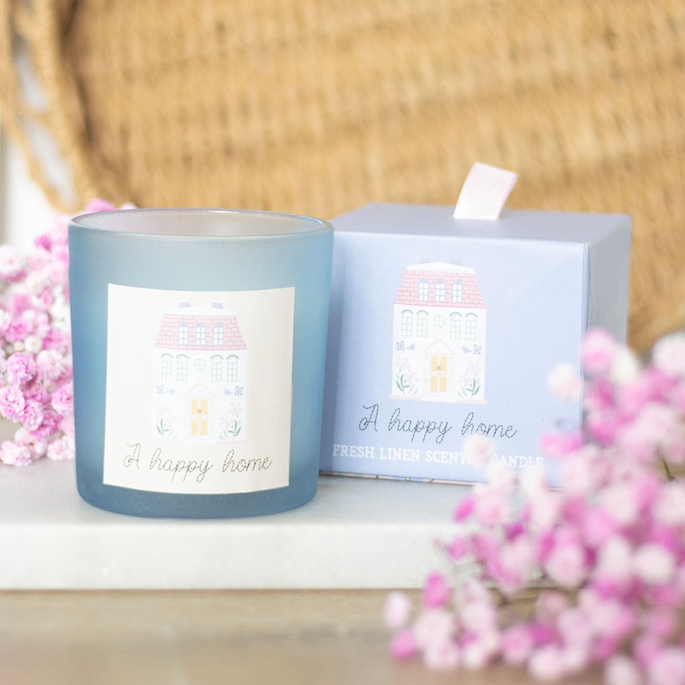 White candle inside blue glass container with a square cream sticker on the front that says 'A Happy Home' in script font at the bottom and a whimsy picture of a house above the text.