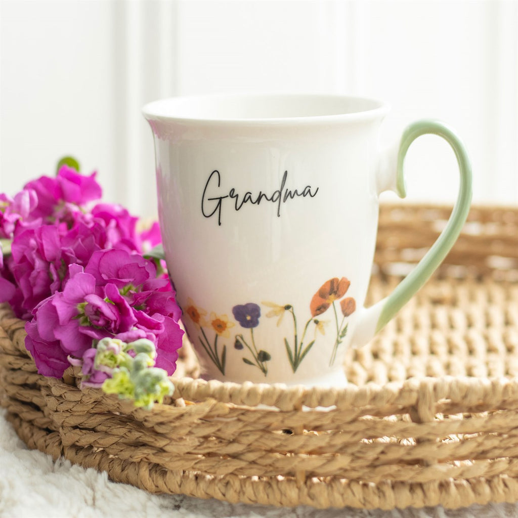 White pedestal china mug with a green handle featuring a floral design at the bottom of the mug, and 'Grandma' written in script font on the middle of the mug. 